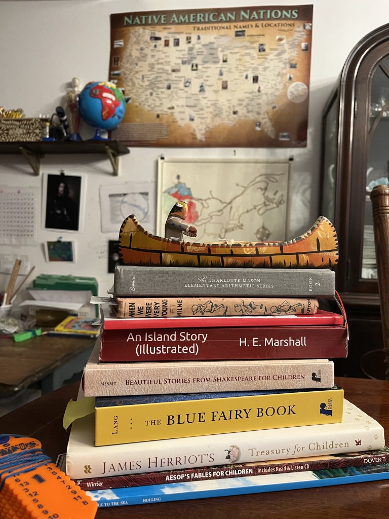 A stack of books from Year 1. On the wall behind is a map of the Great Lakes as well as map of the U.S. with the Native American Nations. Also on the wall is a star chart and artwork. A colorful globe sits on a shelf.