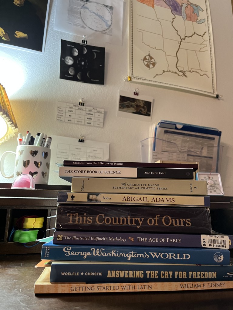 A stack of books from Year 5 on a desk. On the wall above the desk is a map of the Mississippi River, a chart of the lunar cycle, and tables of latin verbs.